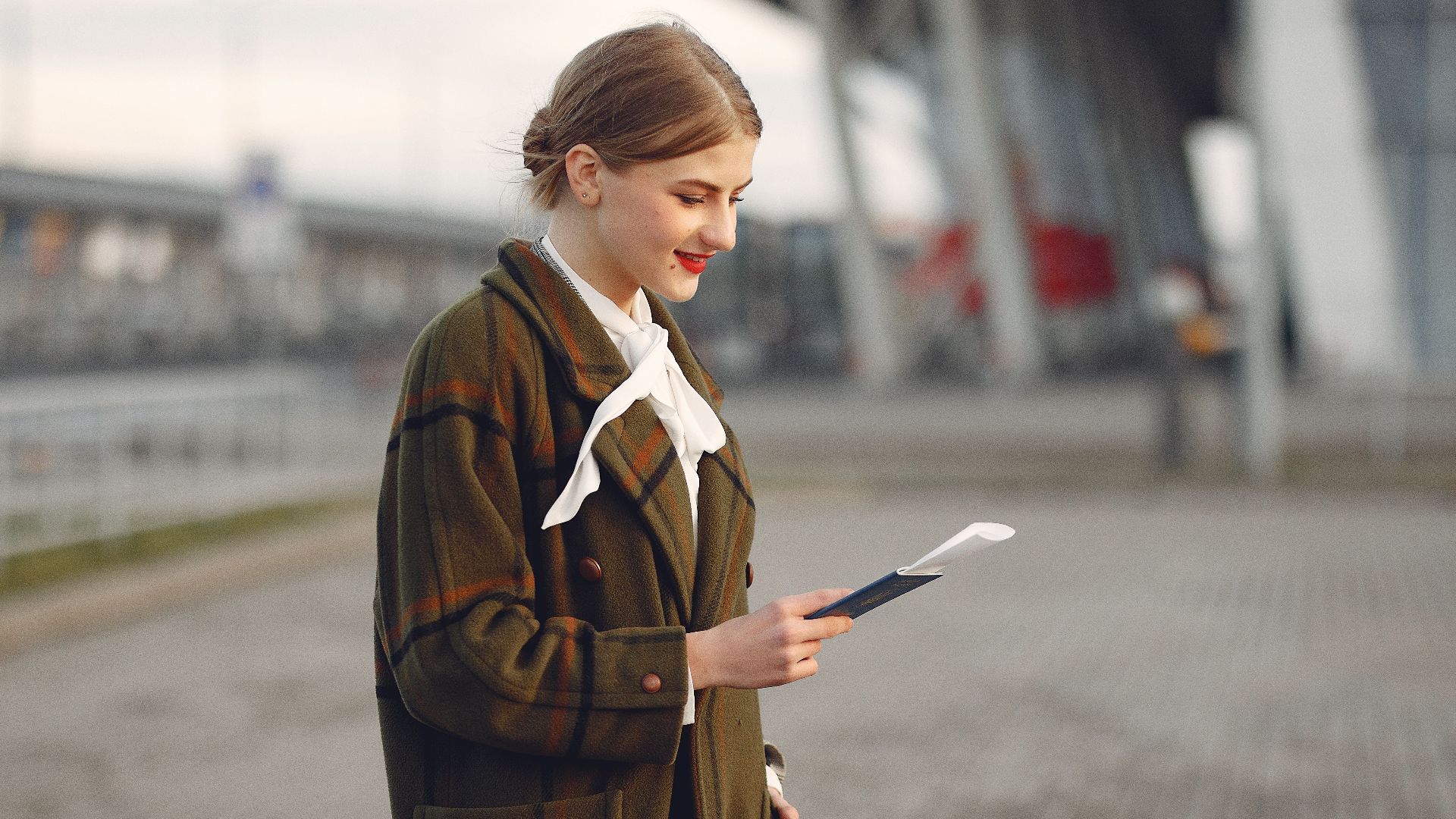 Smiling female passenger wearing trendy plaid coat and white blouse checking passport and ticket standing on pavement near contemporary building of airport