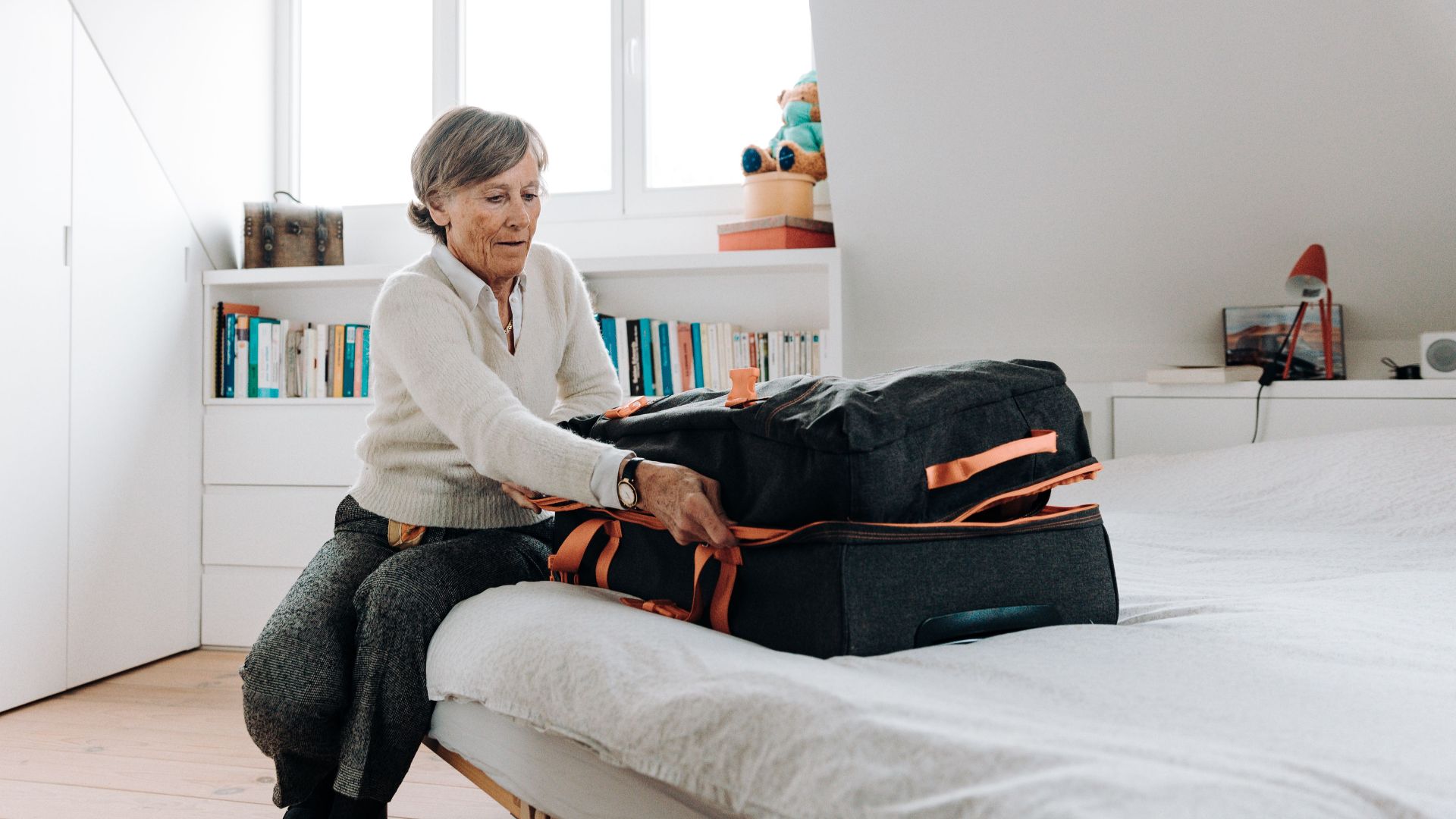 Elderly woman packing suitcase on bed in bright, modern bedroom setting.