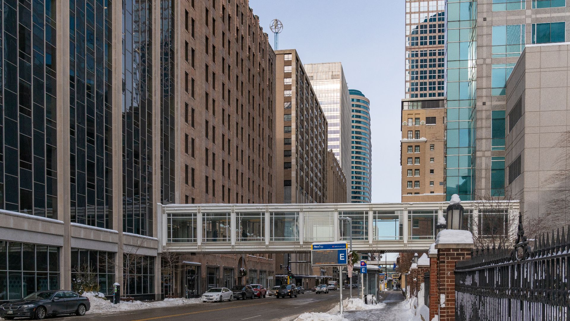 An enclosed skyway or walkway connects buildings in downtown Minneapolis, Minnesota, above a cold city street with shoveled sidewalks in winter.