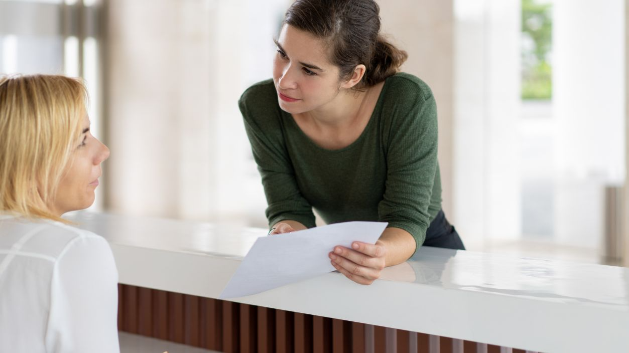 Shutterstock-1169871532, Serious female tourist trying to check in. Girl in casual leaning on reception desk, holding paper and talking to receptionist. Hotel and paperwork concept