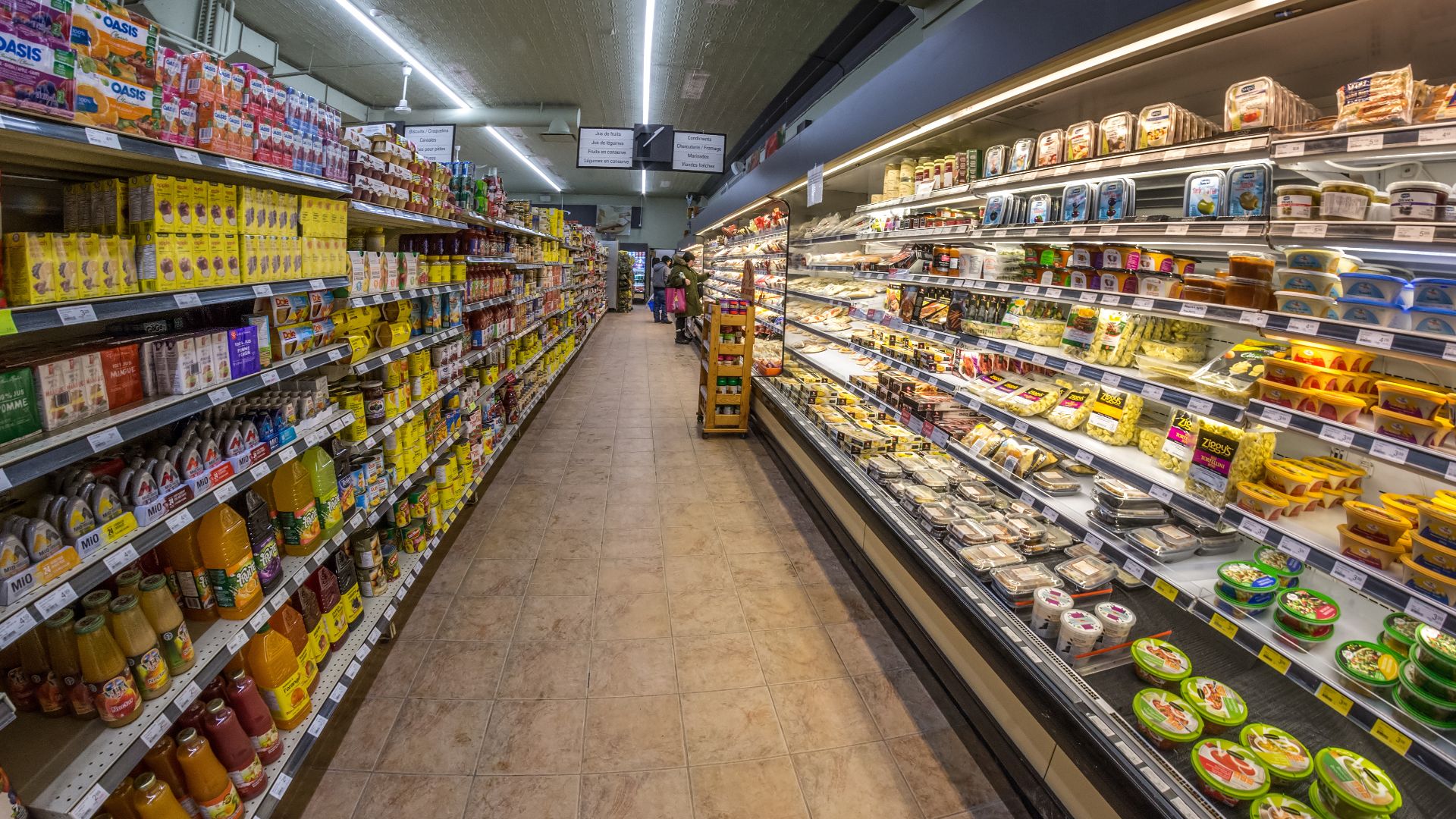 An aisle of the Intermarché supermarket, jars and bottles of preserves on the shelves to the left and the refrigerated cases of cured meat and cheese to the right, a single shopper at the far end. Quebec City, Quebec, Canada. Photographed on Rue Saint-Jea