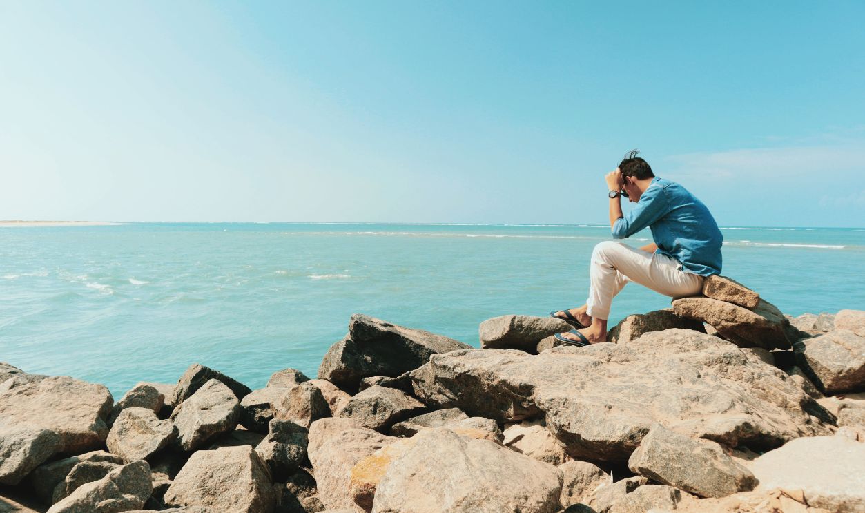 A man in casual attire sitting on rocks by the ocean