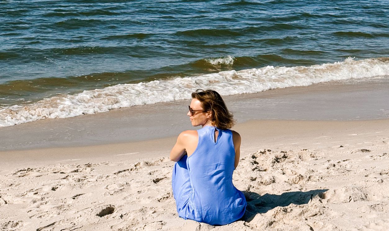 A woman in a blue dress sits on a sandy beach, enjoying a peaceful view of the Baltic