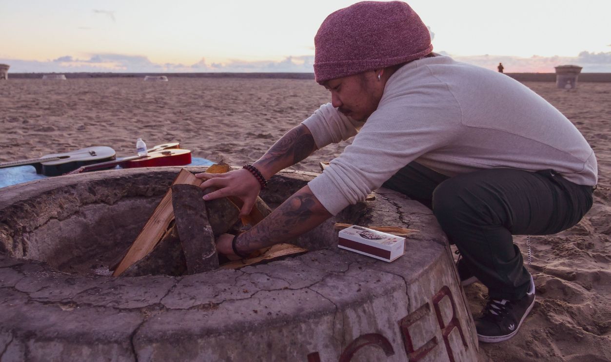 A man prepares a fire pit on a sandy beach with guitars nearby
