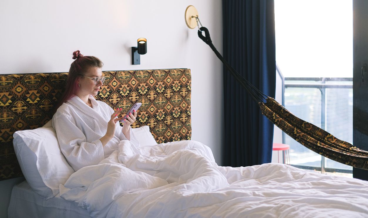 Woman in a bathrobe sits on a bed using a smartphone in a comfortable hotel room.