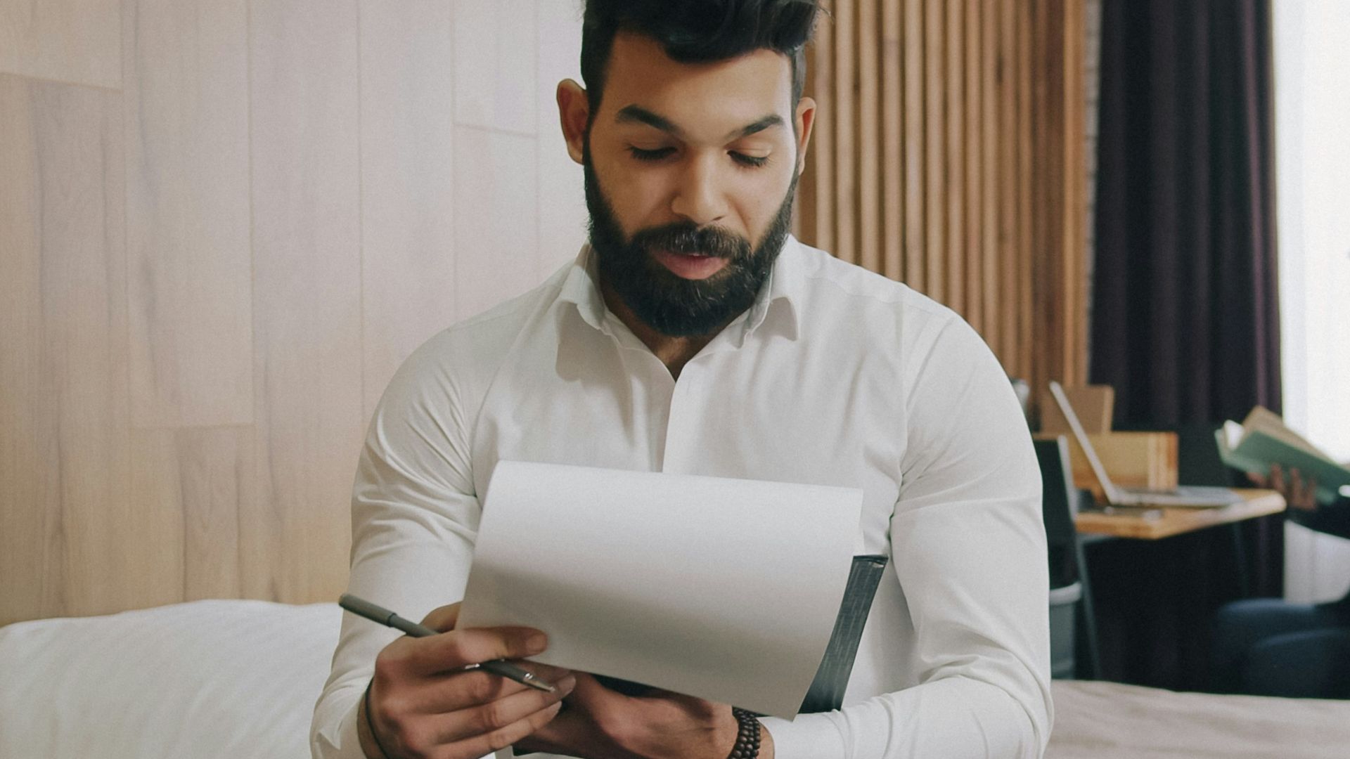 A man in a white shirt writing in a notebook on a hotel bed