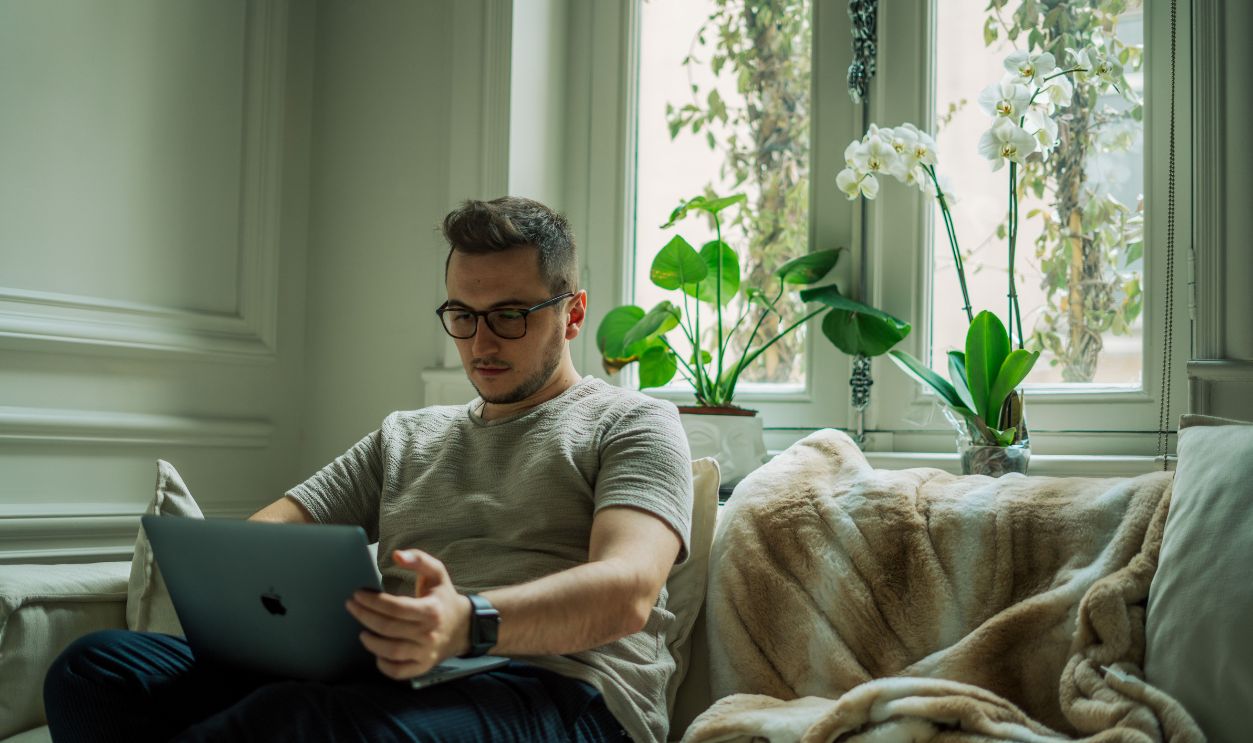A man with glasses works on a laptop while seated on a sofa, surrounded by indoor plants