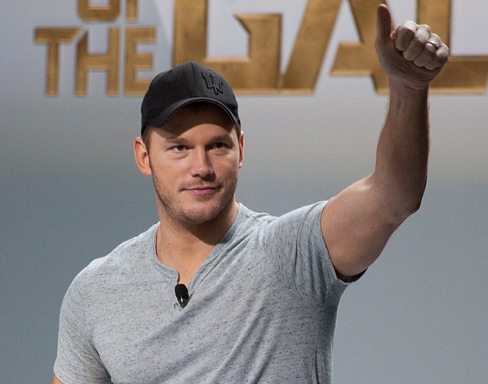 Portrait Photo of Chris Pratt in a gray t-shirt and his arm raised.