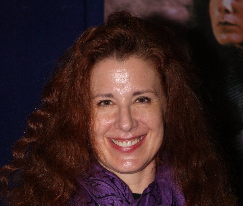 Portrait Photo of Suzie Plakson wit a purple scarf, smiling