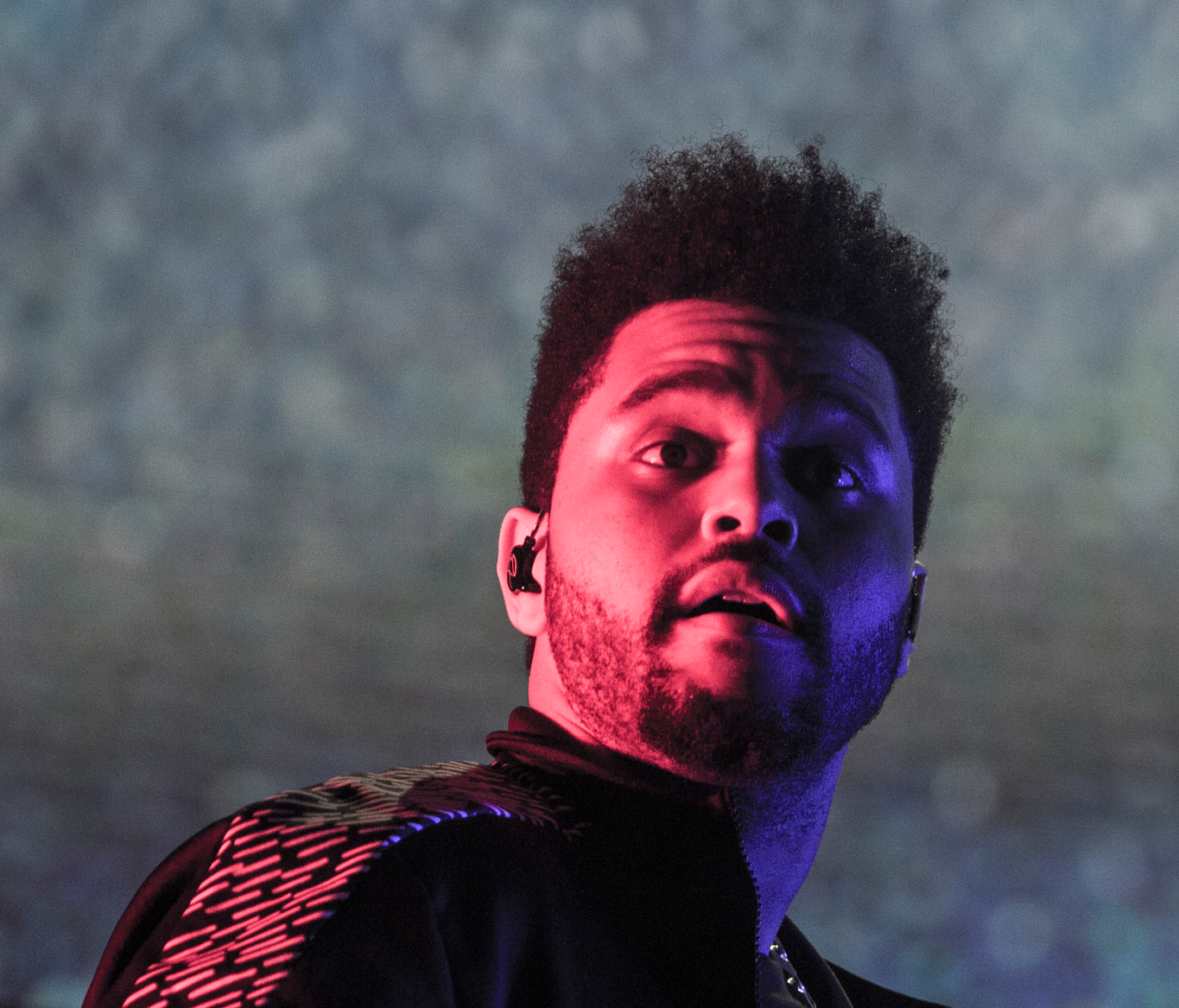 The Weeknd wearing a jacket in pink and blue lighting, holding a microphone