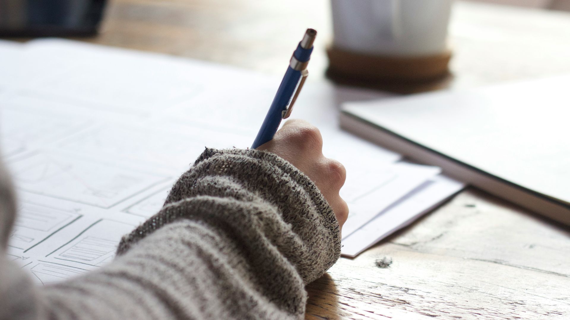 person writing on brown wooden table near white ceramic mug