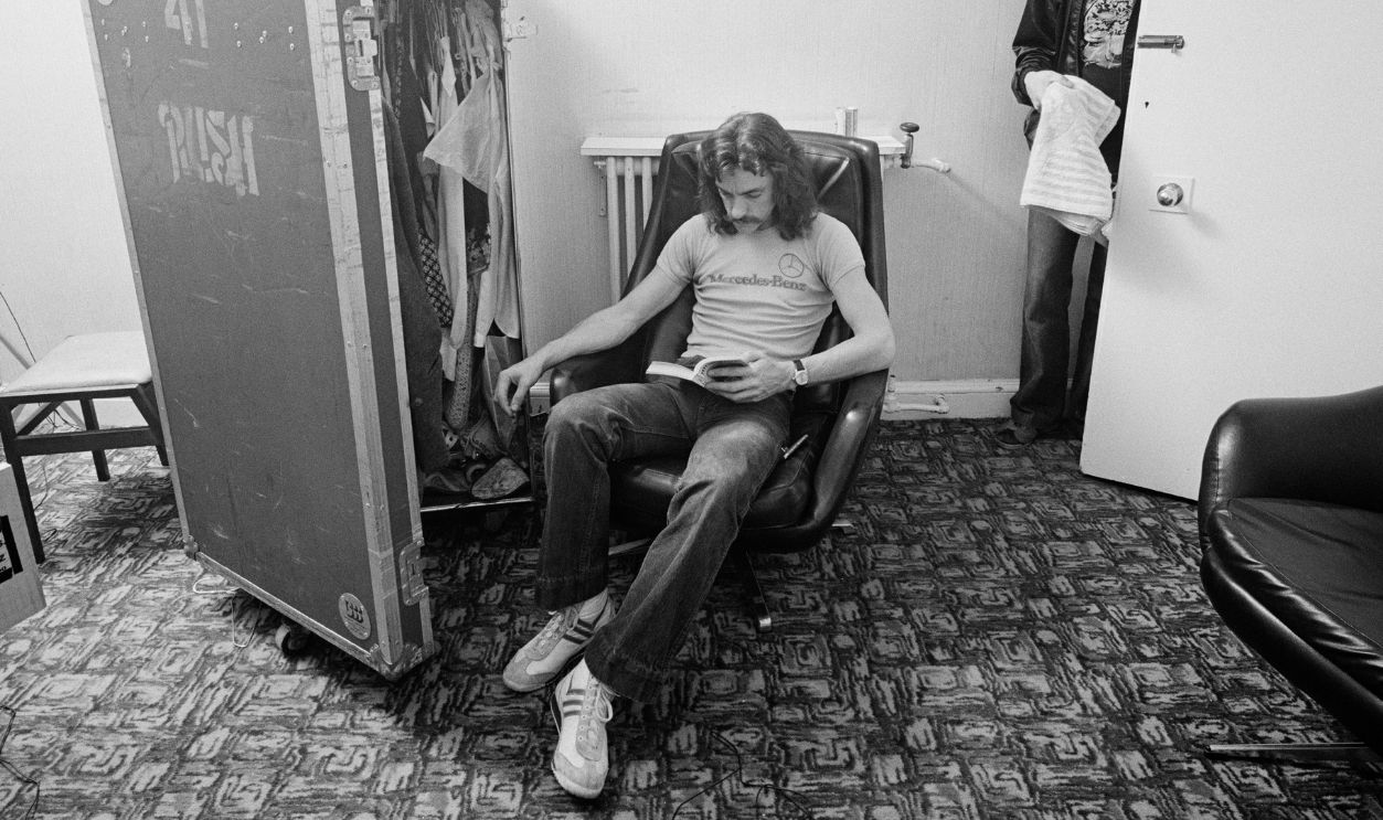 Gettyimages - 123262408, Rush Backstage Neil Peart, drummer with Canadian rock band Rush, sitting backstage in armchair, reading a book beside a travel wardrobe with the door ajar, at the Gaumont in Southampton, Hampshire, England, United Kingdom, 13 May 1979. The door to the room is open, with a man walking through carrying a towel.