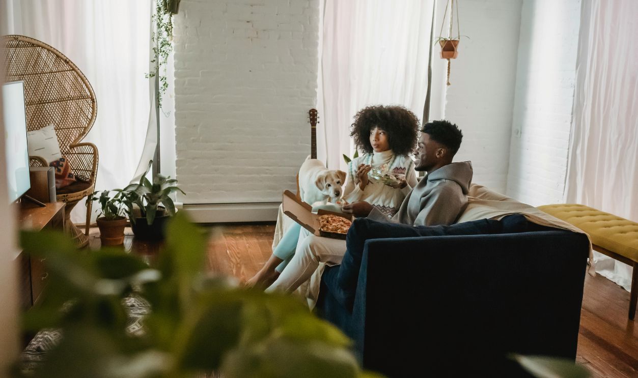 Focused black couple eating pizza and watch TV near dog