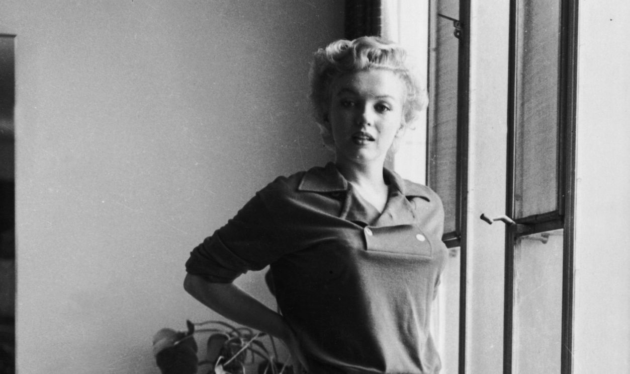 circa 1955: American actor Marilyn Monroe (1926 - 1962) stands barefoot near a window in a sweater and checkered pants.