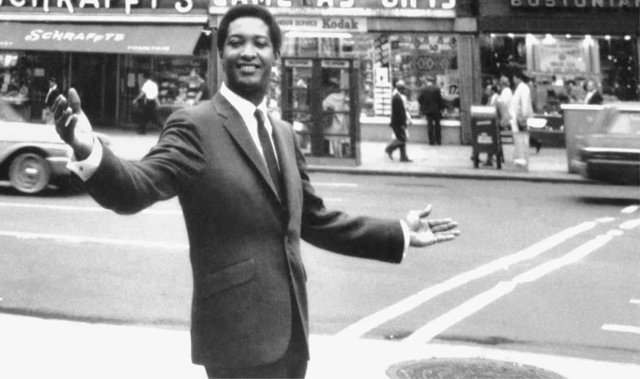 Soul singer Sam Cooke stands on a sidewalk matching his pose on a billboard across the street, New York, New York in 1964.
