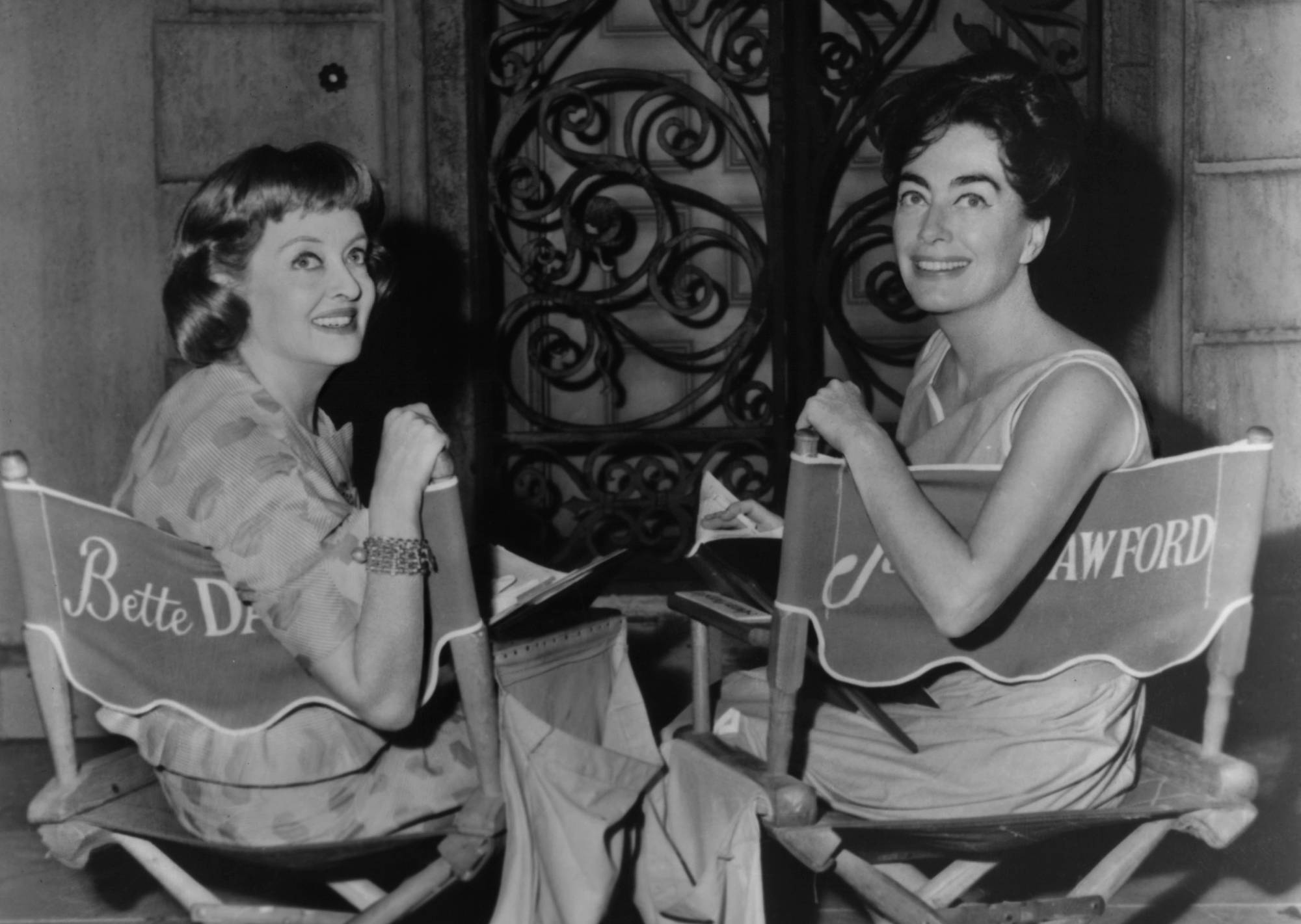 Bette Davis and Joan Crawford  seated on chairs with their names on