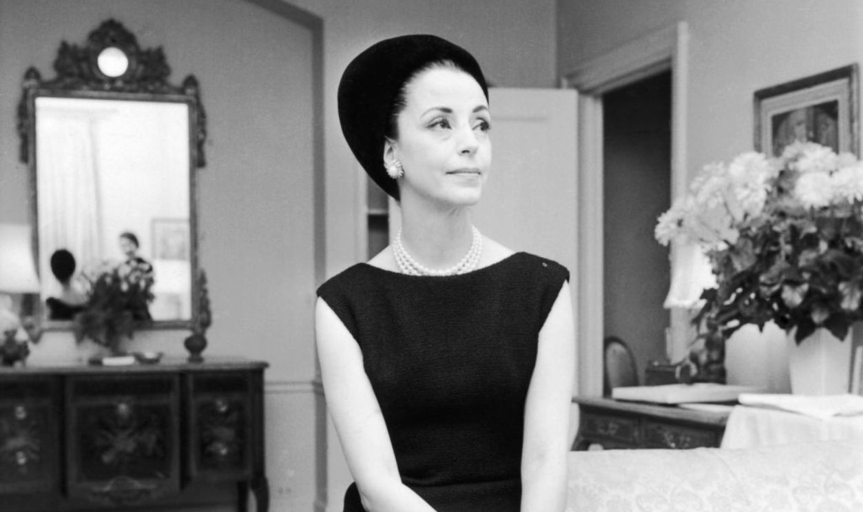 Portrait of fashion muse Gloria Guinness wearing a black capsleeve dress and black velvet Balenciaga hat in her Waldorf Towers suite