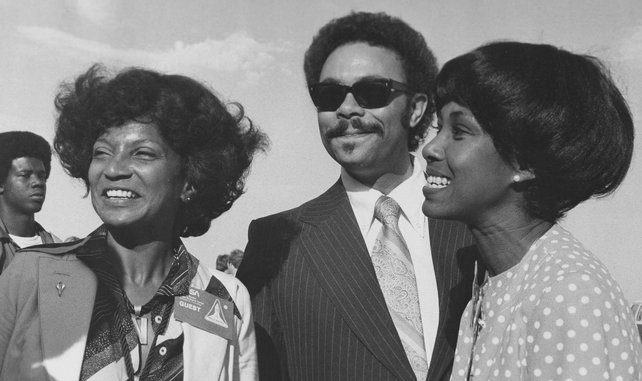 Black and white photograph of, from left, actress Nichelle Nichols, Curtis Graves, Director of Community Affairs at NASA (National Aeronautics and Space Administration), and Congresswoman Yvonne Burke, at the first flight and landing of the Space Shuttle Orbiter Enterprise, August 2, 1977. 