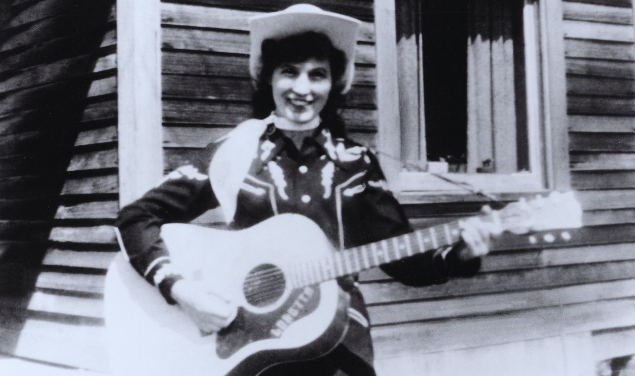 Loretta Lynn Portrait With Acoustic Guitar CIRCA 1960: Loretta Lynn holds her acoustic guitar as she poses for a portrait wearing a cowboy hat, a scarf and western shirt outside a log cabin in circa 1960.