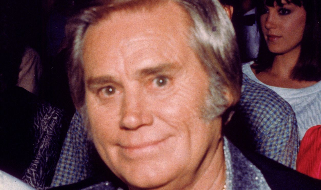 Portrait of American country music vocalist George Jones seated at an unidentified event, late 1980s. He is dressed in a black jacket with a leaf design, a silver lame shirt, and a gold necklace with interlocking letters 'G' and 'J' on it.