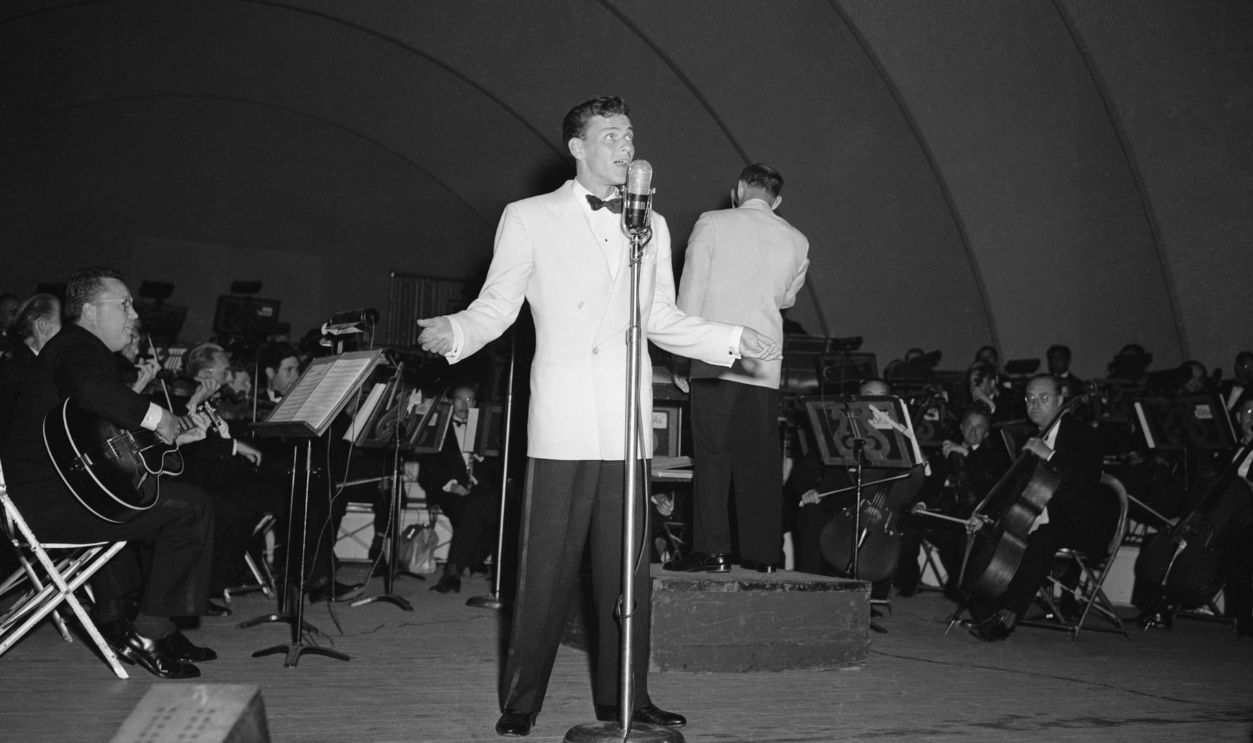 Frank Sinatra Crooning Frank Sinatra performs at the Hollywood Bowl. Here he stands full length crooning into a microphone, wearing a white jacket and black pants.