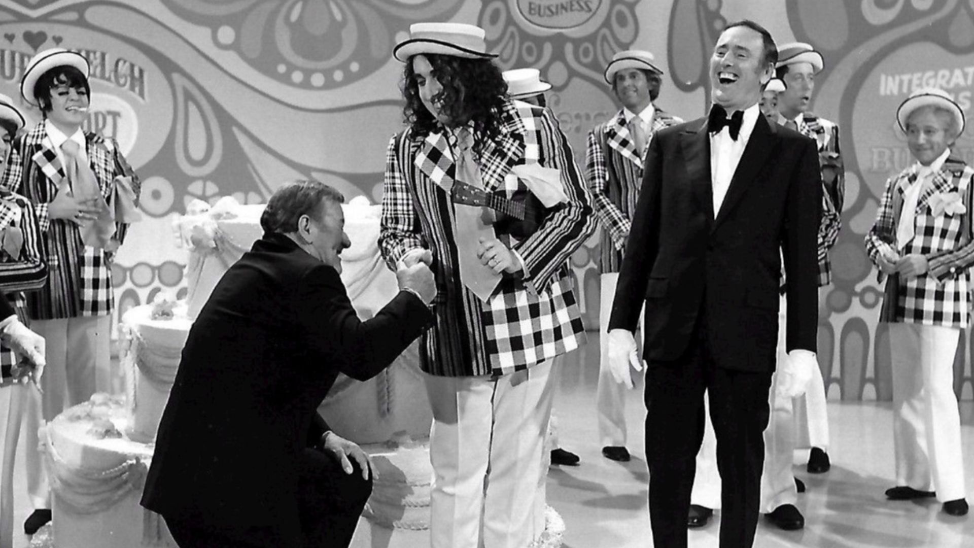 Publicity photo from the 100th episode of the television program Rowan & Martin's Laugh-In.  Guest stars are John Wayne and Tiny Tim. Host Dick Martin is pictured, along with past and present cast members from the show.  Visible in the background are Ruth