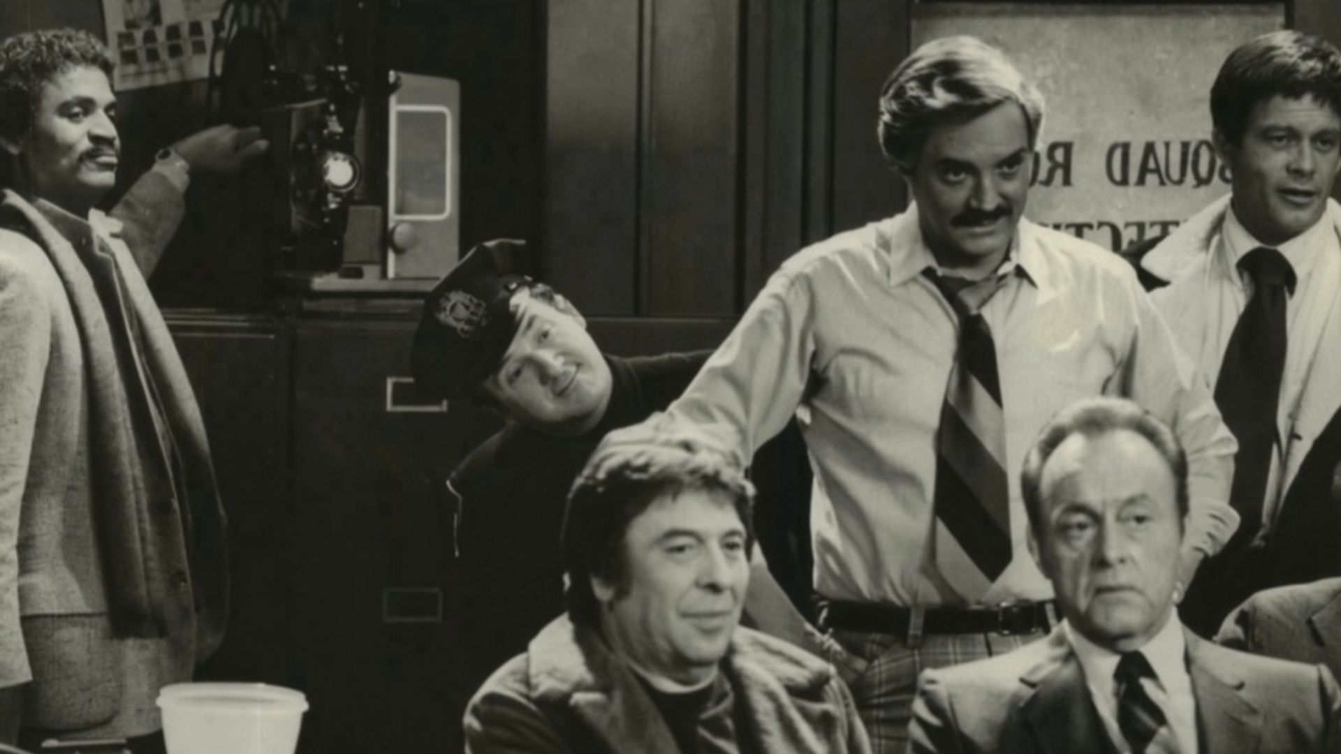 A very proud Det. Harris (Ron Glass, left) flicks the switch on the projector as he is about to premiere the adult movie he has written, directed and produced. His audience includes his fellow detectives plus criminal suspects and their victims, in