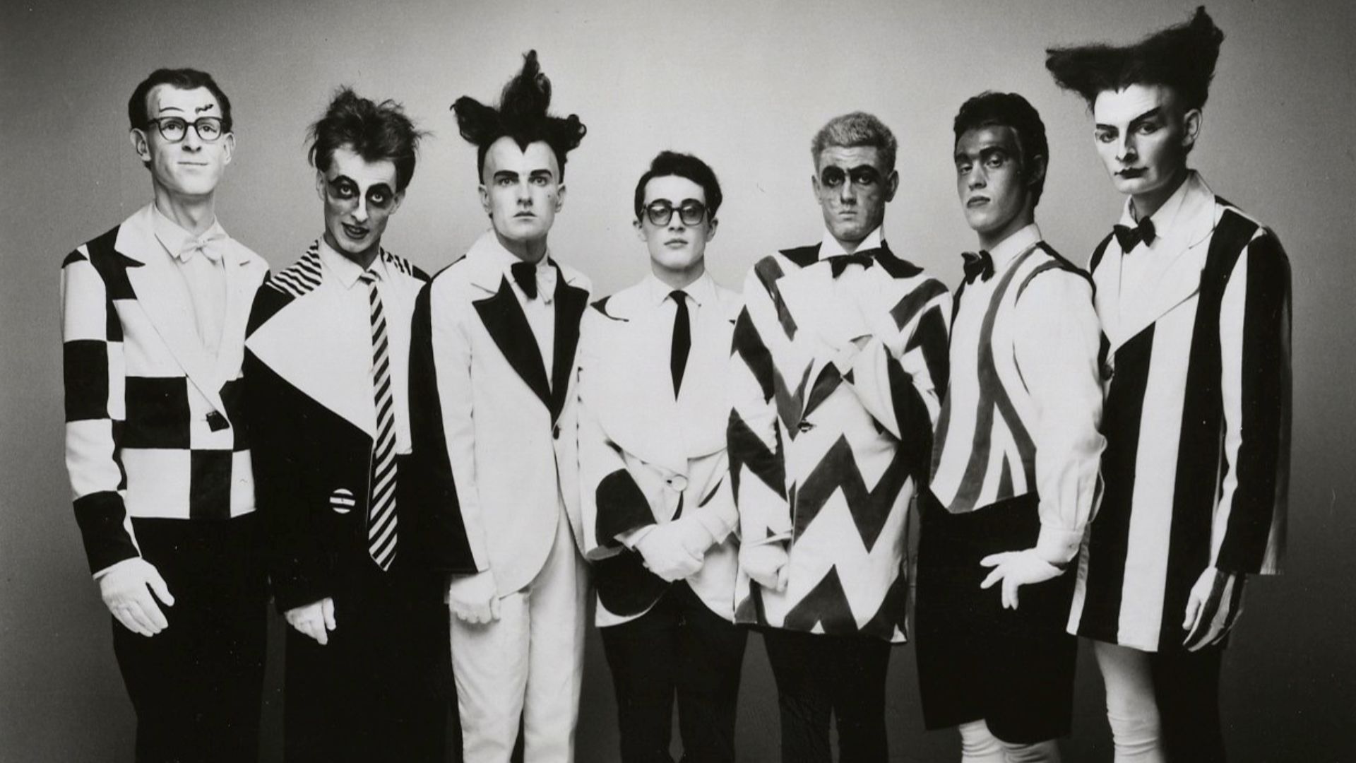 Seven men with theatrical dress and make-up standing and looking at the camera, black and white. Under photo is the text