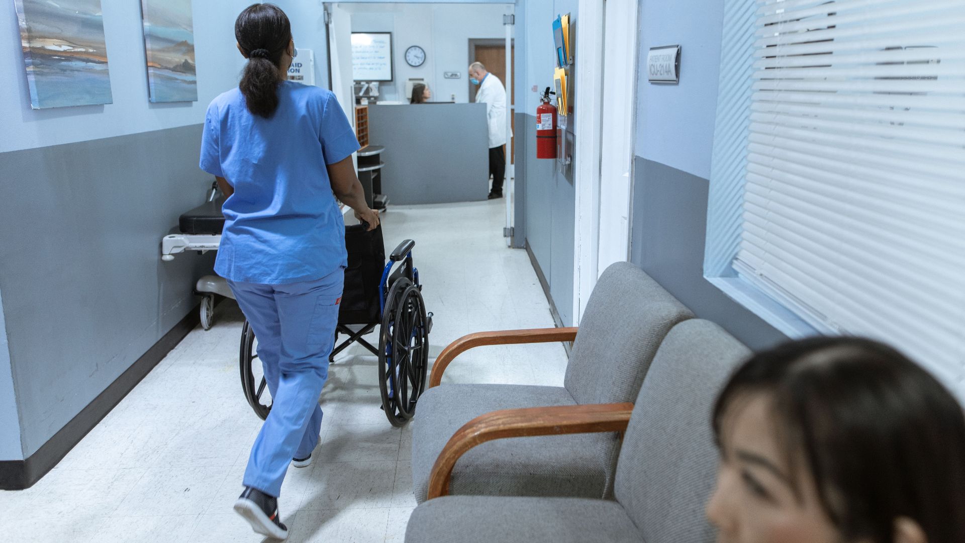 A nurse in scrubs pushes a wheelchair through a well-lit hospital corridor, attending to patient care.