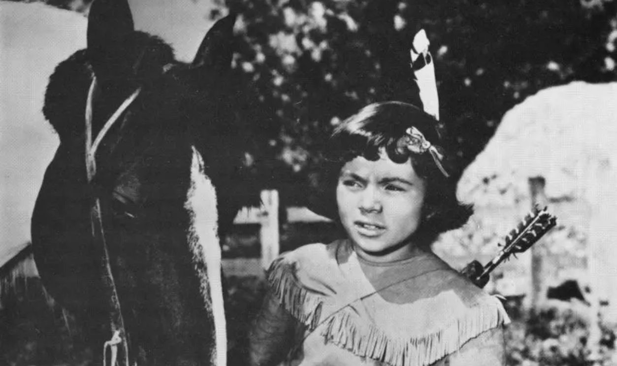 American child actor Robert Blake stands outdoors beside a horse, wearing a fringed cowhide poncho and a feather in his hair as the character 'Little Beaver' in a still from one of the 'Red Ryder' Western film series, circa 1945.