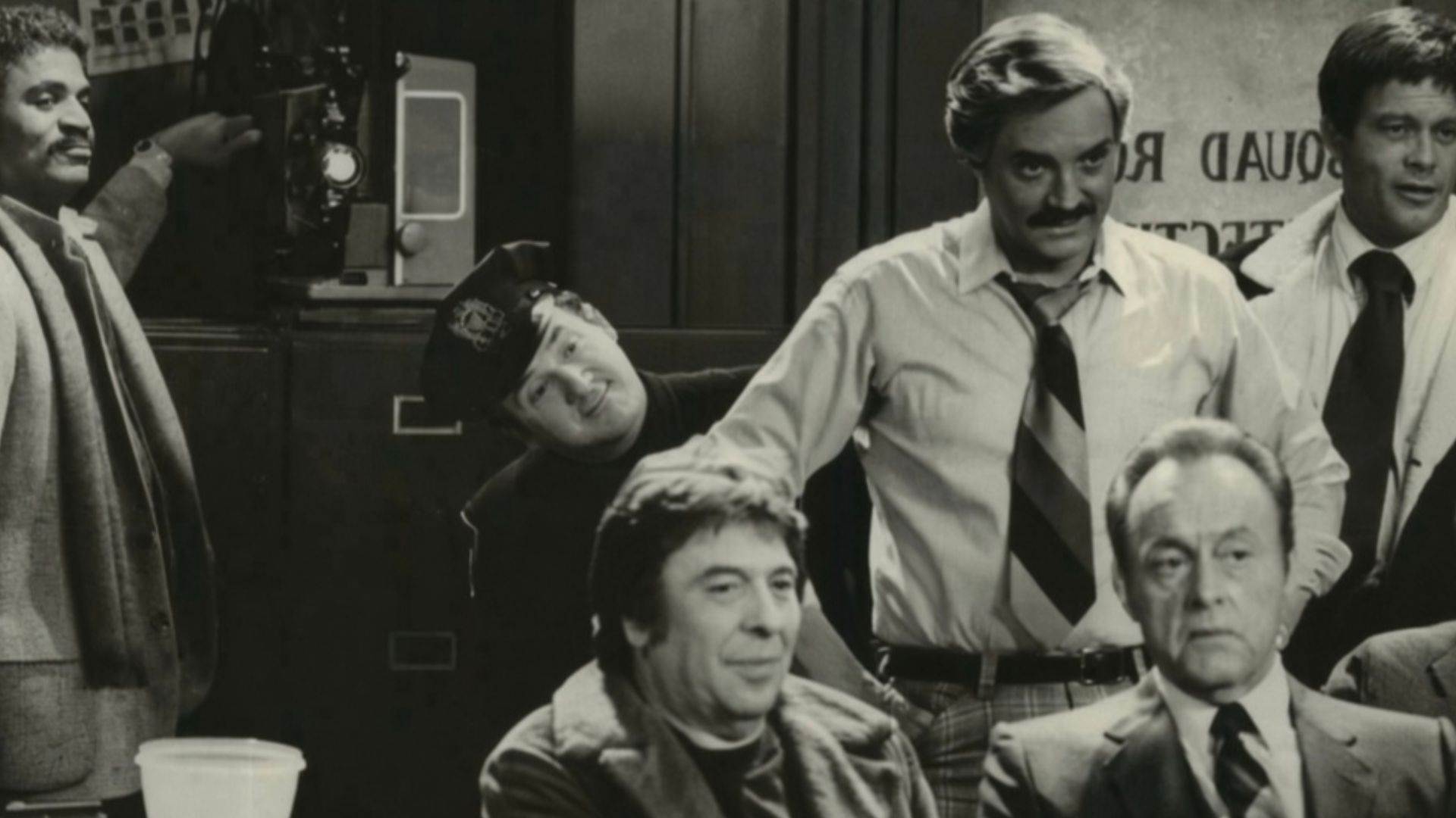 A very proud Det. Harris (Ron Glass, left) flicks the switch on the projector as he is about to premiere the adult movie he has written, directed and produced. His audience includes his fellow detectives plus criminal suspects and their victims, in