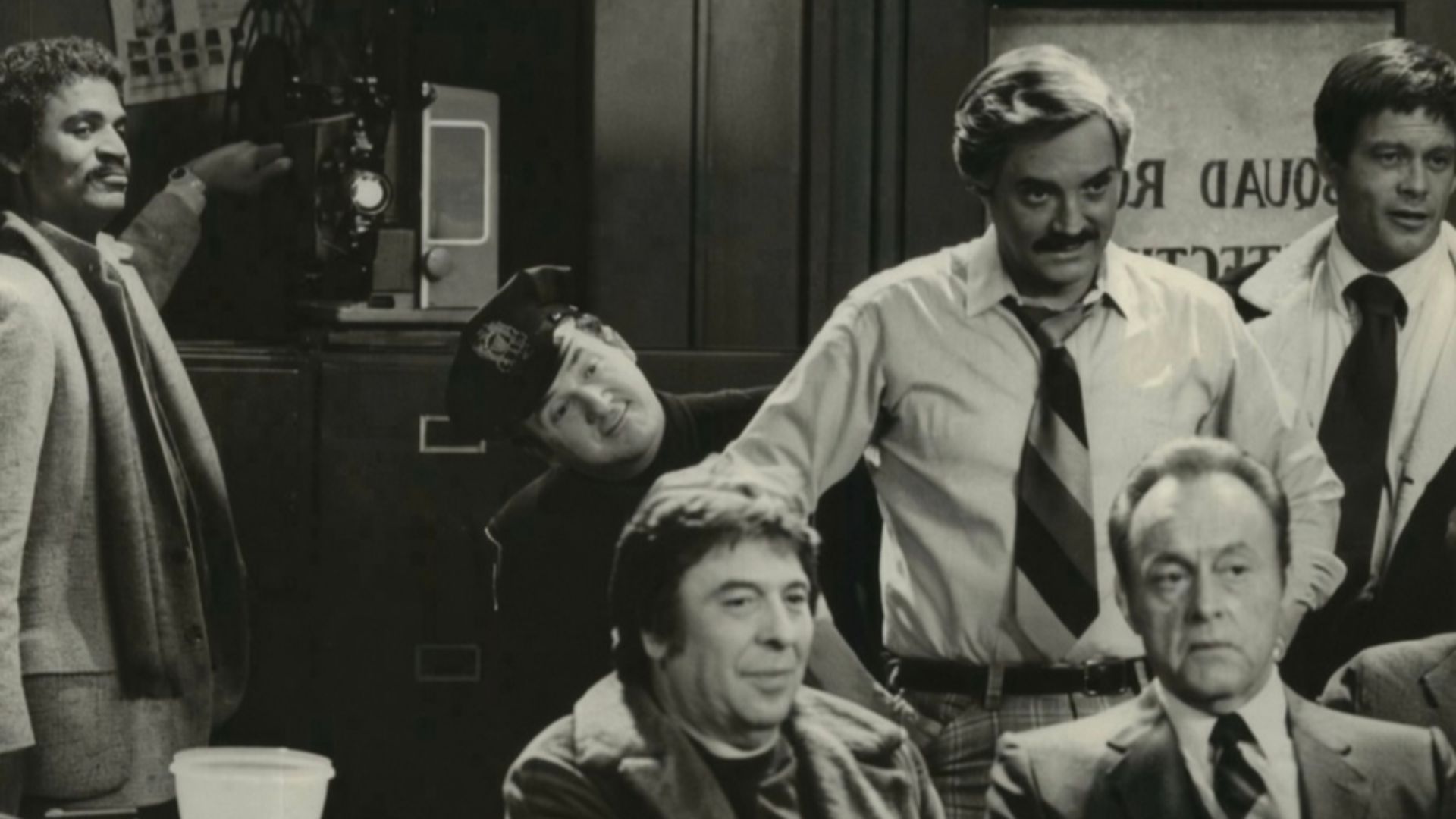 A very proud Det. Harris (Ron Glass, left) flicks the switch on the projector as he is about to premiere the adult movie he has written, directed and produced. His audience includes his fellow detectives plus criminal suspects and their victims, in