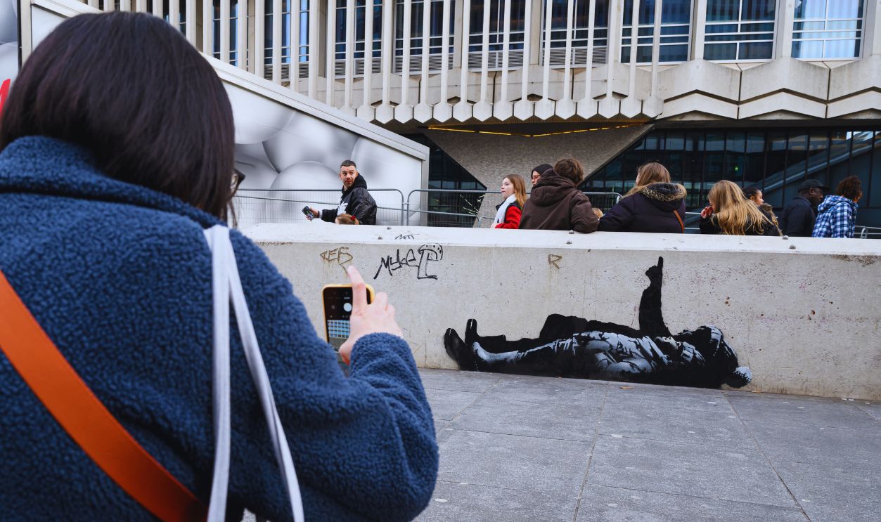 Gettyimages - 2253129121, New Banksy Artworks Appear In London LONDON, ENGLAND - DECEMBER 22: A woman stops ot take a photo of an apparent Banksy artwork which has appeared on a wall near to the Centrepoint building on December 22, 2025 in London, England. This afternoon, the renowned street artist Bansky shared a post on Instagram indicating authorship of an identical work that appeared in another part of London. 