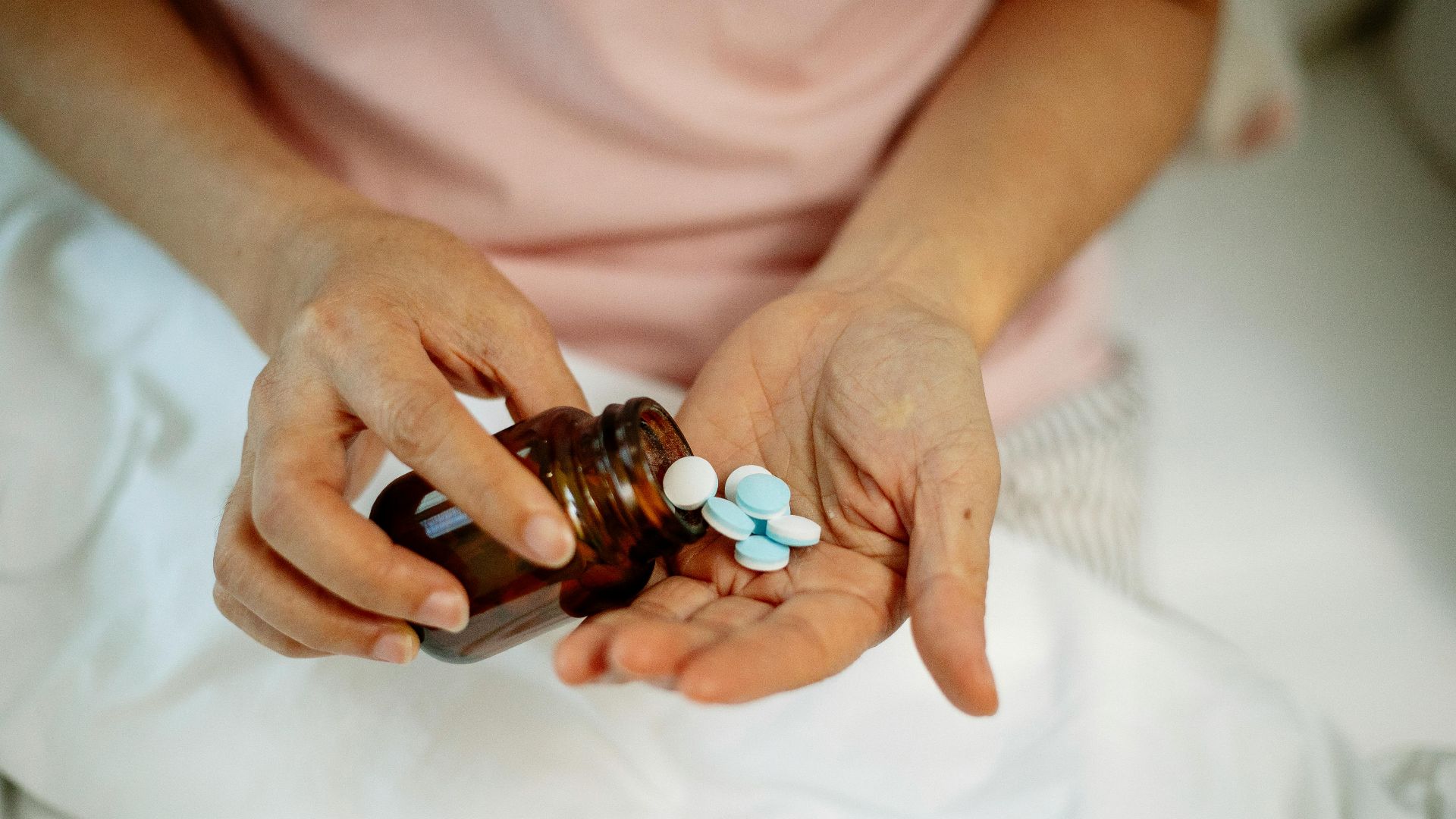 An anonymous woman in bed pouring blue and white pills from a brown bottle into her hand.