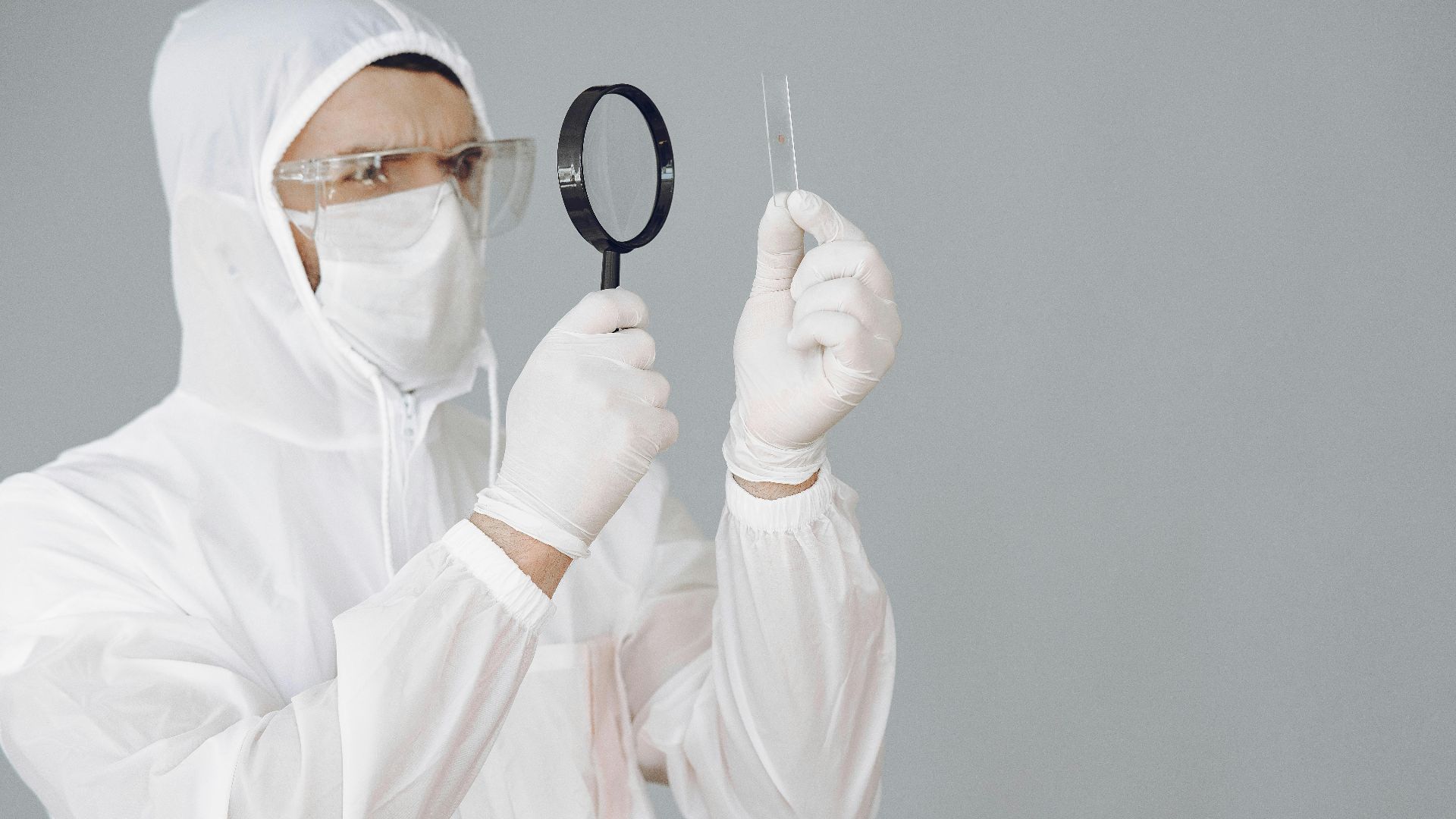 A scientist in protective gear examines a sample slide with a magnifying glass in a laboratory setting.