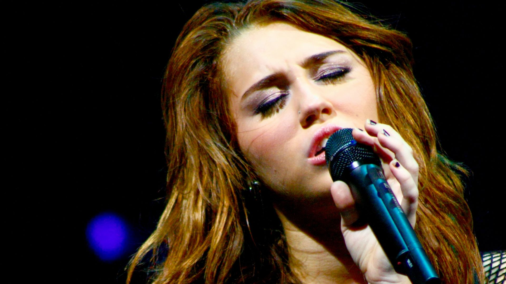 Miley Cyrus, a brunette teenager, sings with a microphone close to her in a concert that is part of her Wonder World Tour in Auburn Hills.