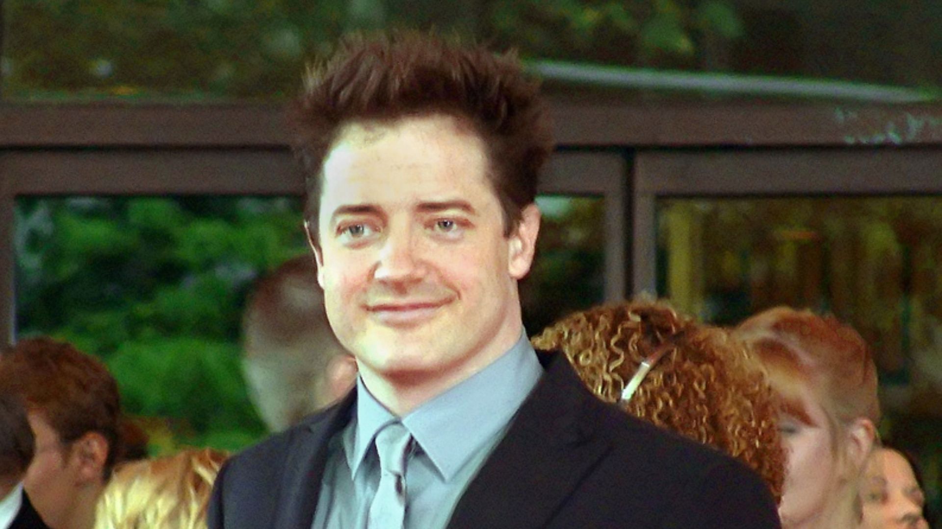 La photographie montre l'acteur américain Brendan Fraser en plan rapproché. L'acteur a la peau claire, les yeux bleu-gris, les cheveux noirs. Il sourit au public situé hors de la photo. Il porte un costume sombre et, en-dessous, une chemise et une cravate gris perle.