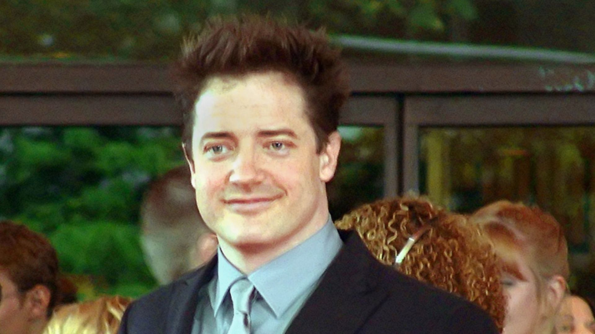 La photographie montre l'acteur américain Brendan Fraser en plan rapproché. L'acteur a la peau claire, les yeux bleu-gris, les cheveux noirs. Il sourit au public situé hors de la photo. Il porte un costume sombre et, en-dessous, une chemise et une cravate gris perle.