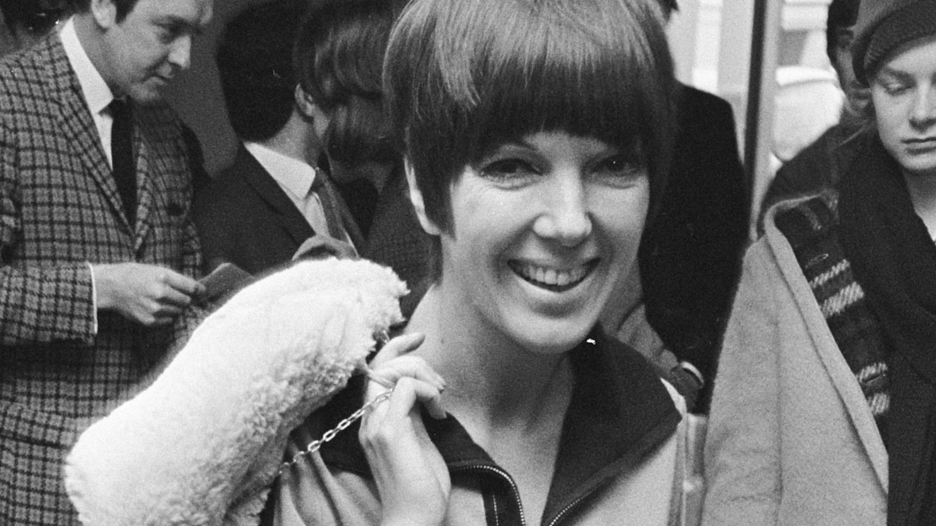 Mary Quant wearing a mini dress of her own design, with a sheepskin coat and bag thrown over her shoulder, and wearing go-go boots.