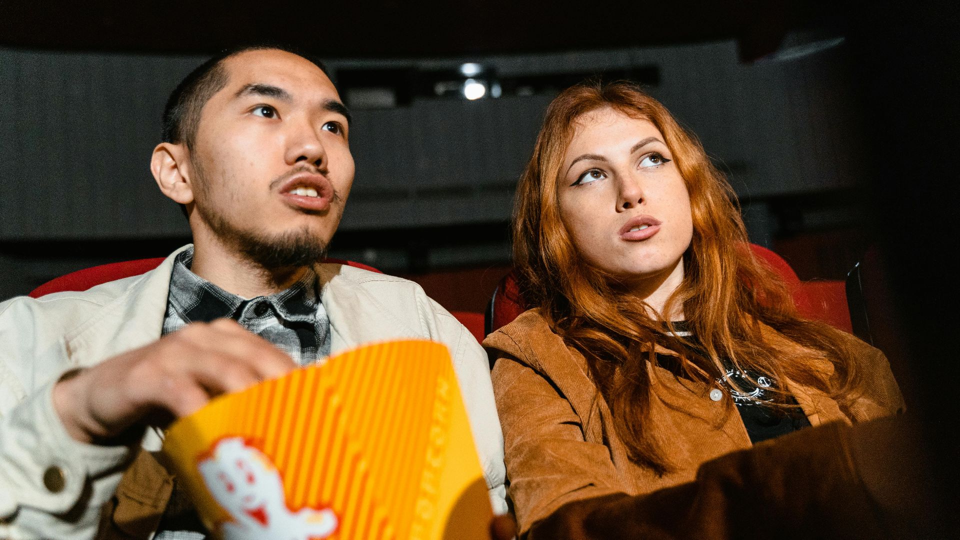 A couple sits in a movie theater, engrossed in a film while enjoying popcorn.