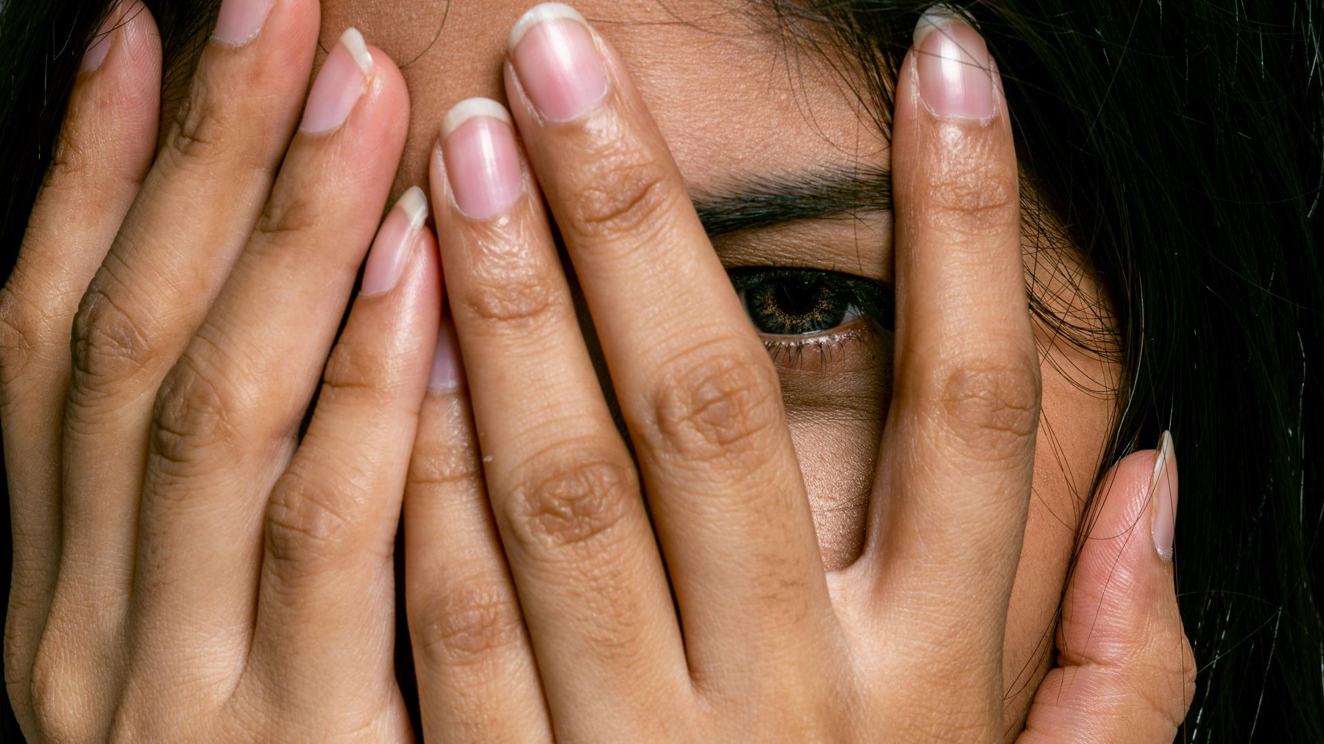 Close-up of woman covering part of her face with hands, revealing one eye.