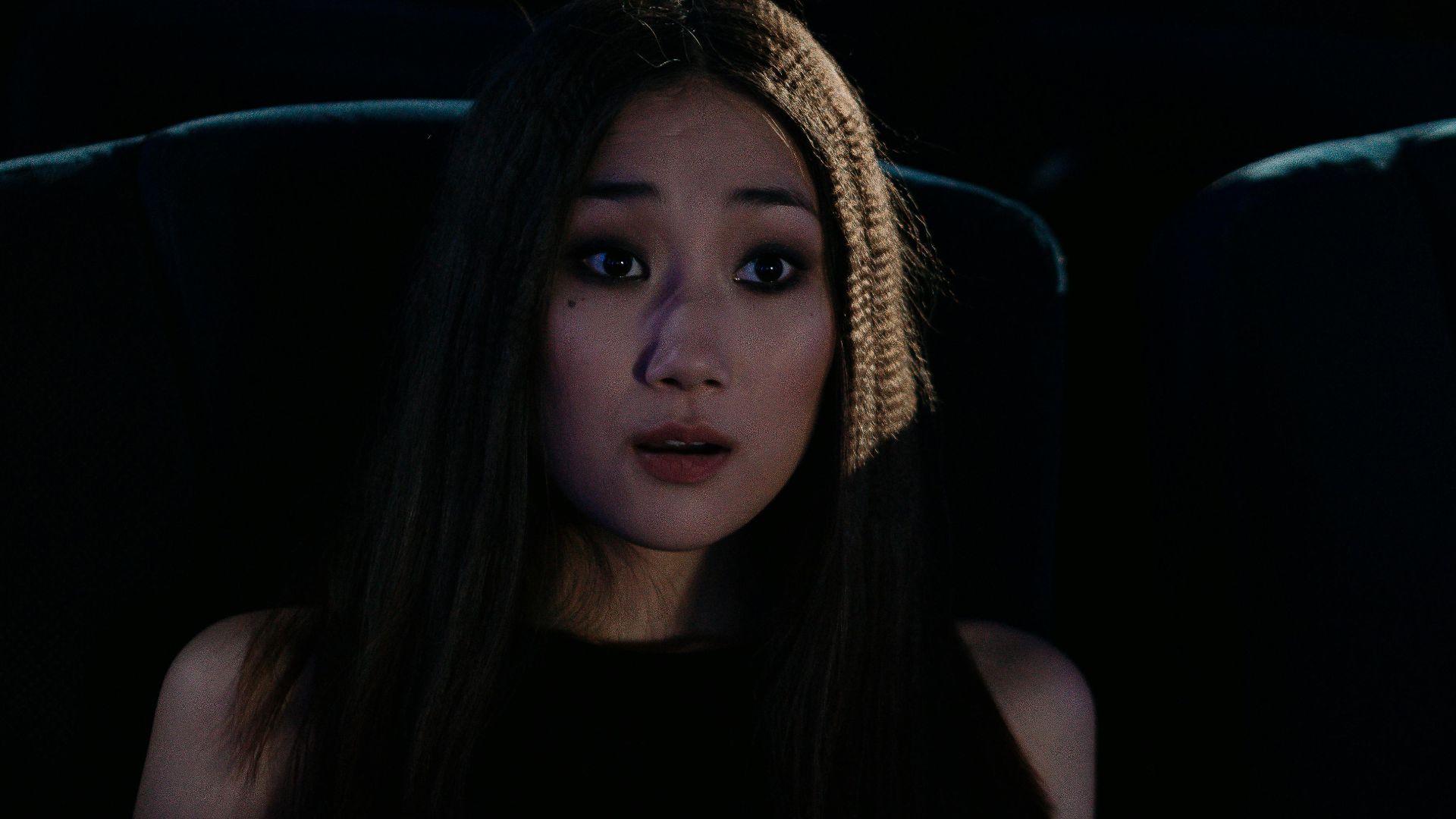 Asian woman holding popcorn looking surprised in a dark cinema.