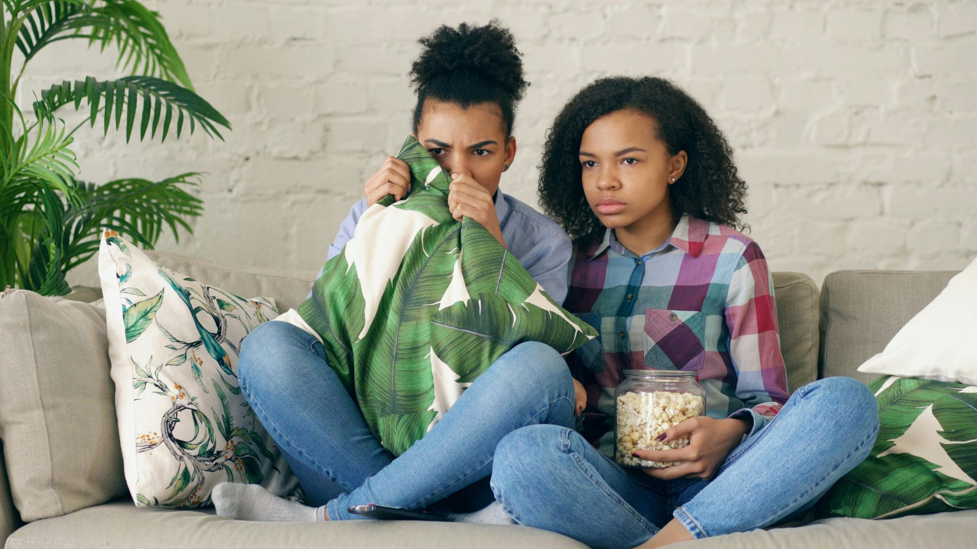 Two young girls sitting on a couch watching a scary movie with popcorn.