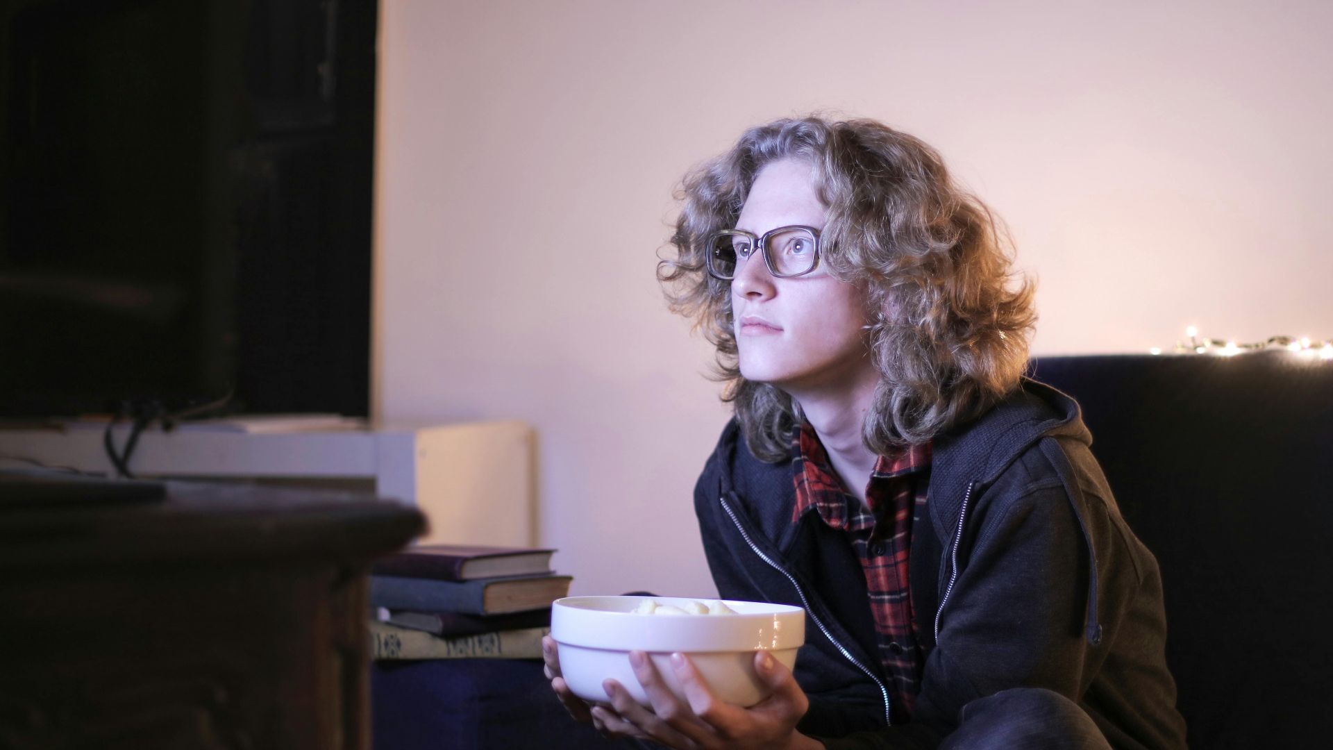 Serious young male with long curly hair sitting with crossed legs on couch in living room and enjoying interesting movie while chilling at home during weekend