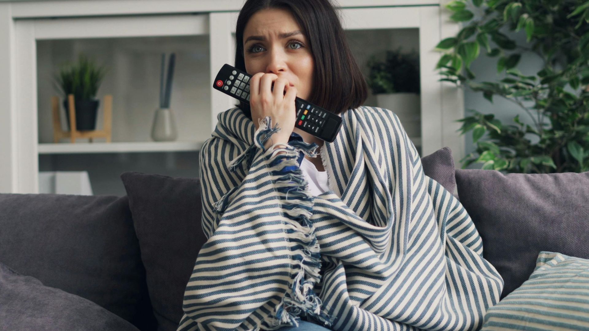 Anxious woman wrapped in a blanket watching TV at home with a remote in hand.