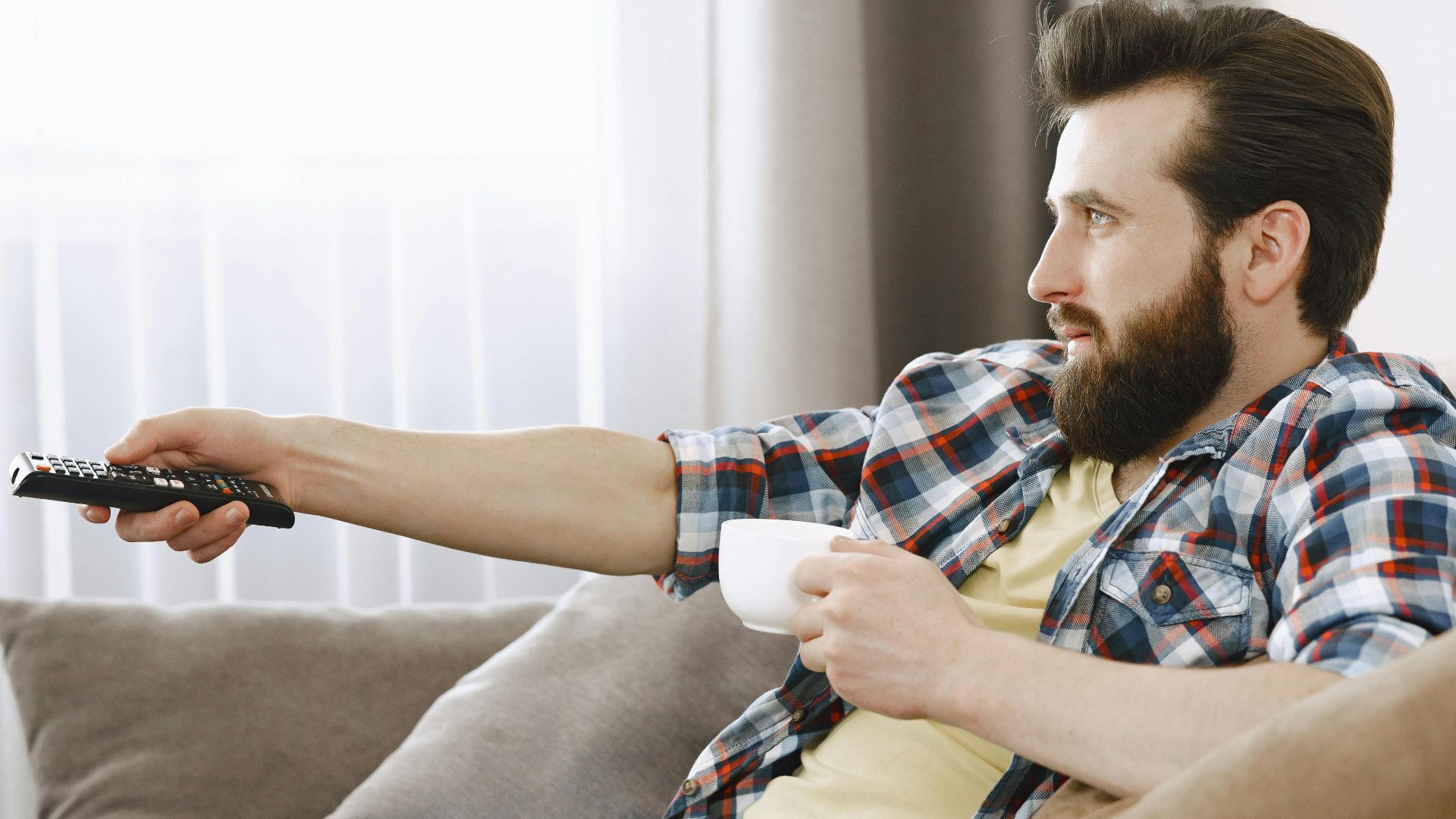 Bearded man in casual wear relaxing on a sofa, enjoying coffee while watching TV indoors.