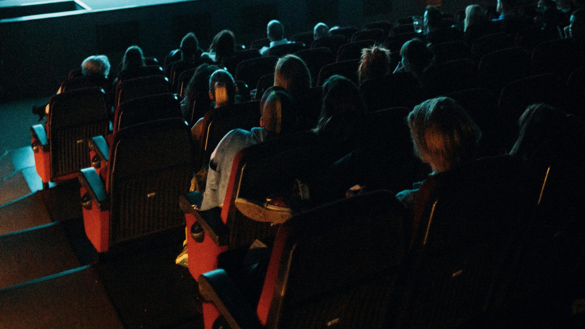 Crowded cinema theater with audience watching a movie on a large screen in a dark setting.
