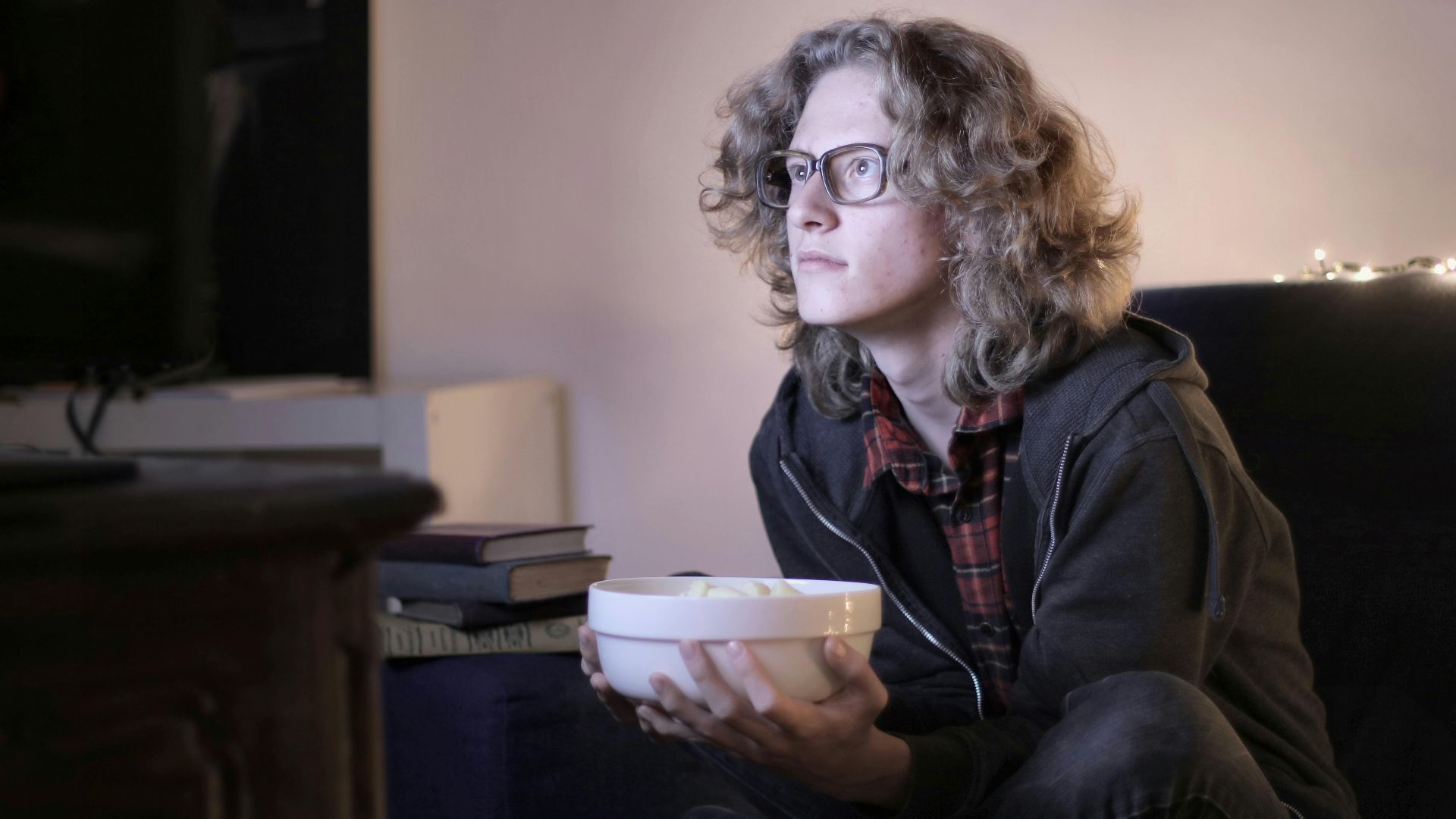 Concentrated male teenage with long hair in casual wear and eyeglasses sitting on couch with bowl of popcorn and watching interesting movie while resting in cozy living room at home