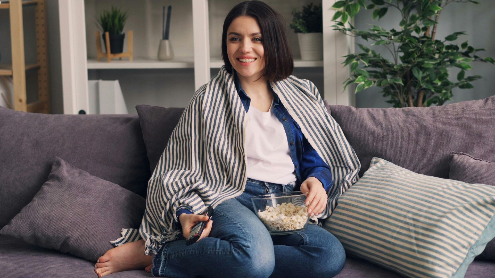 Smiling woman sitting on couch with popcorn, enjoying a cozy indoor moment.