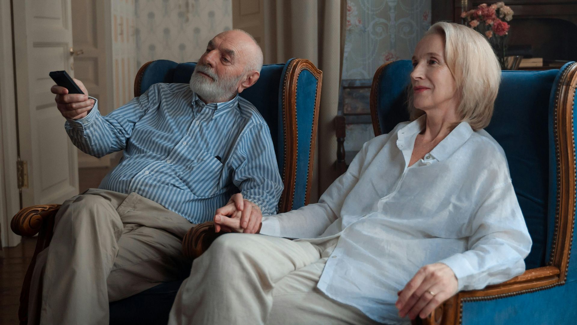 An elderly couple sits holding hands, enjoying a peaceful moment at home.